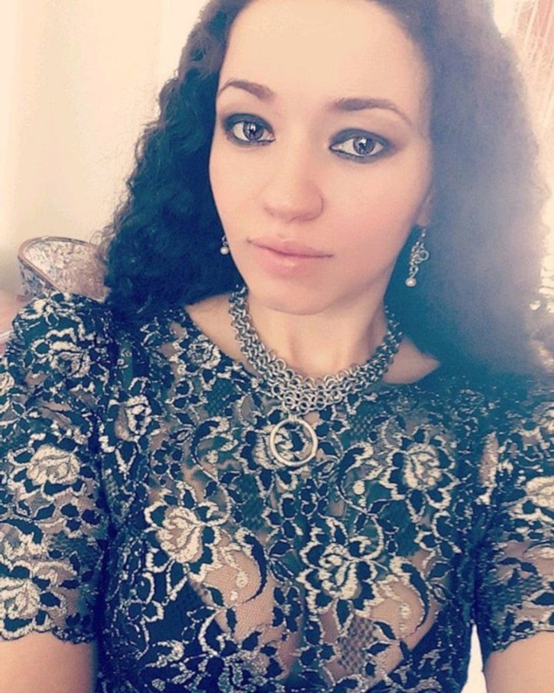 May include: A woman wearing a black lace top with a silver chain necklace and silver earrings. The necklace has a large silver ring pendant.