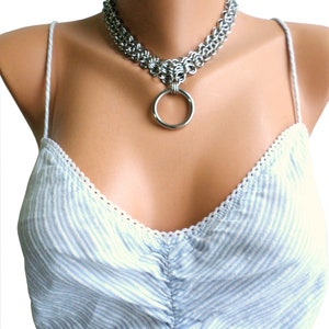 May include: A silver chainmail choker necklace with a large silver ring pendant. The necklace is worn on a mannequin with a light blue and white striped top.