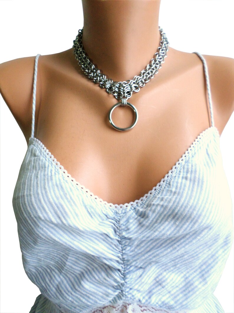 May include: A silver chainmail choker necklace with a large silver ring pendant. The necklace is worn on a mannequin with a light blue and white striped top.