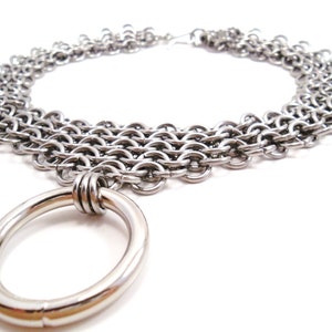 May include: A silver chain necklace with multiple rows of interlocking rings and a large, smooth, silver ring pendant.