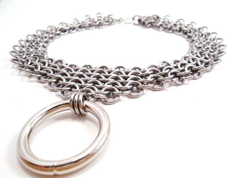 May include: A silver chain necklace with multiple rows of interlocking rings and a large, smooth, silver ring pendant.