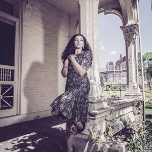 May include: A woman in a black lace dress stands on a porch with a white railing. She is looking off to the side and has her hands clasped in front of her. The porch is decorated with ornate details and there is a building in the background.