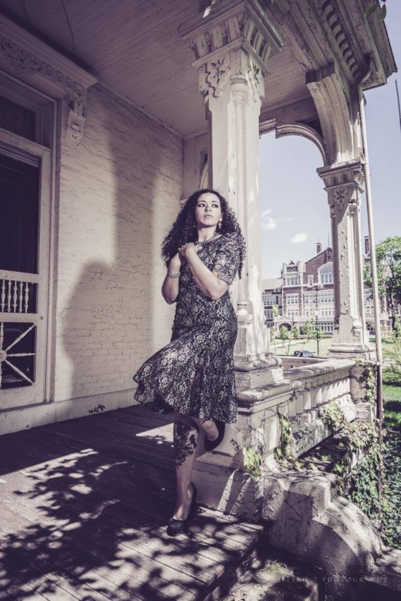 May include: A woman in a black lace dress stands on a porch with a white railing. She is looking off to the side and has her hands clasped in front of her. The porch is decorated with ornate details and there is a building in the background.