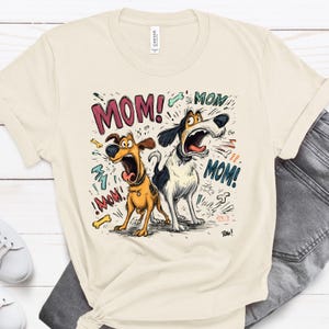 May include: Cream-colored t-shirt featuring a cartoon illustration of two dogs with open mouths, shouting "MOM!". The design includes bone accents and speech bubbles. The shirt is paired with denim jeans and white sneakers.
