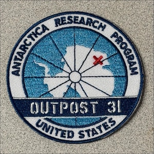 May include: A circular patch with the text "ANTARCTICA RESEARCH PROGRAM" and "OUTPOST 31 UNITED STATES". The patch features a map of Antarctica in white and blue, with a red cross marking a location.