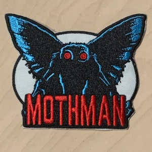 May include: A black and blue embroidered patch with a mothman creature with red eyes and the word "MOTHMAN" in red lettering.