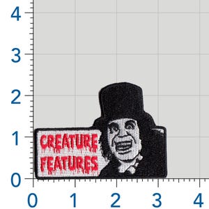 May include: An embroidered patch featuring a black and white portrait of a person in a top hat next to a white rectangular sign with the red text "CREATURE FEATURES" in a dripping font. The patch measures approximately 3.5 inches by 2.25 inches.