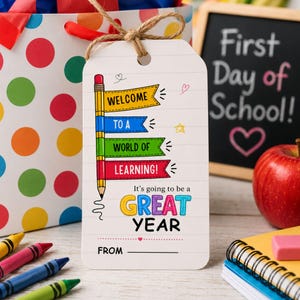Könnte beinhalten: Ein weißes, rechteckiges Geschenkanhänger-Schild mit einer Bleistift-Illustration und bunten Bannern mit der Aufschrift Welcome to a world of learning! It's going to be a great year. Der Anhänger hat eine Juteschnur und eine Zeile für einen Namen nach From.