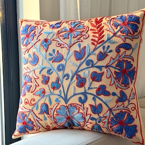 Può includere: Cuscino quadrato beige con ricami floreali intricati nei toni del blu e del rosso. Il design ricamato presenta fiori e foglie stilizzati, creando un motivo decorativo. Il cuscino ha un bordo color crema.
