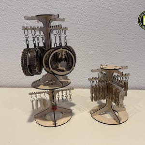 Könnte beinhalten: Zwei Schlüsselanhänger-Ausstellungsständer aus Holz. Der größere Ständer hat drei Etagen mit Schlüsselanhängern, die von jeder Etage hängen. Der kleinere Ständer hat zwei Etagen mit Schlüsselanhängern. Die Schlüsselanhänger haben verschiedene Formen und Designs. Die Ständer sind aus hellem Holz.