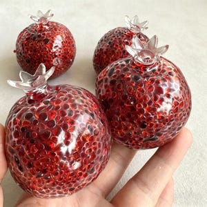 May include: Four decorative glass pomegranates. The pomegranates are a deep red color with a textured surface, and topped with a clear, crown-like detail. The pomegranates are a unique home decor item.