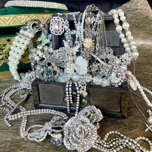 Puede incluir: Un cofre del tesoro de madera rebosante de varias joyas. La colección incluye collares, pulseras y pendientes, con perlas, pedrería y detalles coloridos. Una diadema enjoyada se encuentra encima de una caja verde y dorada.