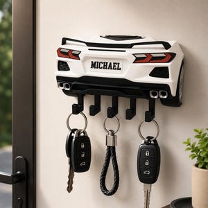 May include: A white wall-mounted key holder shaped like the rear of a sports car, featuring a black spoiler, taillights, and four exhaust pipes. The license plate area displays the name MICHAEL. Five black hooks hold three sets of keys.