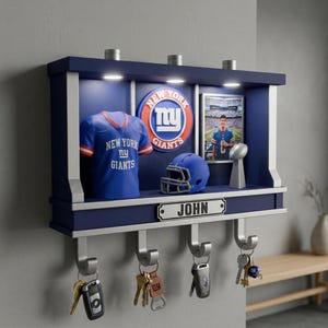 May include: A navy blue wall-mounted display shelf featuring the New York Giants logo, a miniature jersey, a football helmet, a trophy, and a framed photo. The shelf includes three integrated LED lights and four silver hooks holding various keys.