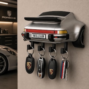 Op de afbeelding: Een zilverkleurige wand-sleutelhouder in de vorm van de achterkant van een sportwagen, met een zwarte spoiler, rode en gele achterlichten en een nummerplaat met de tekst WILLIAM. Vier zwarte haken houden diverse sleutels met Porsche-logo.
