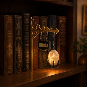May include: A decorative wall-mounted bracket with an ornate gold scrollwork design. Two metal chains hang from the bracket, one holding a black rectangular sign with the name Olivia in gold lettering, and the other holding a glowing cracked egg lamp.