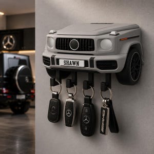 May include: A grey wall-mounted key holder shaped like the front end of a Mercedes-Benz G-Class vehicle. The number plate displays the name SHAWN. Four black hooks extend from the bottom, each holding a car key fob with a silver logo.