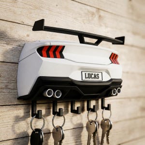 Pode incluir: Um suporte de parede para chaves em forma de traseira de carro esportivo, com um aerofólio preto, lanternas traseiras vermelhas e pretas e uma placa com o nome LUCAS. Inclui quatro escapamentos prateados e cinco ganchos pretos para chaves.