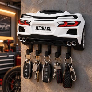 May include: A white wall-mounted key holder shaped like the rear of a sports car, featuring a black spoiler, red taillights, and four exhaust pipes. The number plate area displays the name MICHAEL in black text, with five black hooks holding various keys.