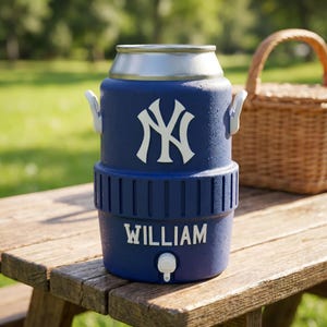 May include: A navy blue insulated drinks cooler with a silver lid and white handles. The cooler displays the New York Yankees logo and the name "WILLIAM" in white. It has a tap for dispensing drinks and is on a wooden picnic table.