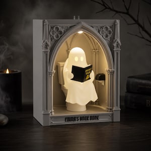 May include: A light-up diorama featuring a white ghost sitting on a toilet inside a gray gothic-style archway. The ghost holds a black book titled Spooky Tales. The base of the structure is inscribed with the text Emma's Book Nook in black letters.