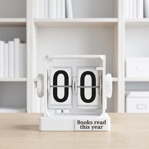 May include: A white flip calendar showing "00" with the text "Books read this year" underneath. The calendar is on a wooden surface, with a white bookshelf and books in the background.