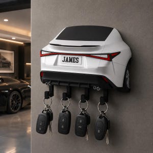May include: A white wall-mounted key holder shaped like the rear of a car, featuring a black roof, red taillights, and a number plate with the name JAMES. Four black hooks extend from the bottom, each holding a black car key fob.