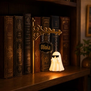 May include: A decorative wall-mounted bracket with an ornate gold-colored scroll design. Hanging from the bracket by metal chains are a rectangular black sign with the name Olivia in gold lettering and a glowing white ghost figure wearing sunglasses.