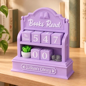 May include: A lavender-colored plastic book tracker display featuring the text "Books Read" at the top and "William's Library" at the base. It includes two shelves with rotatable blocks displaying the numbers 547 and 2600, a small green succulent in a white pot, and a gray cat figurine.
