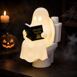 May include: A white, glowing ghost-shaped lamp sitting on a white toilet. The ghost has two oval eye cutouts and holds a small black book with the gold text 'SPOOKY TALES' and a small ghost icon printed on the cover. A roll of toilet paper sits on the tank.