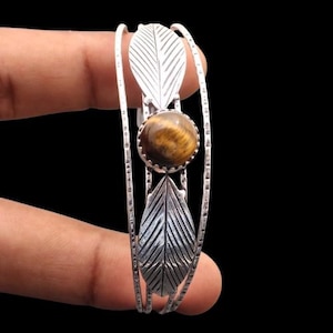 Puede incluir: Un brazalete plateado con una piedra ojo de tigre redonda en el centro, flanqueada por dos adornos en forma de hoja. El brazalete tiene una banda texturizada y se muestra sobre un fondo negro. La piedra mide aproximadamente 2 cm de diámetro.