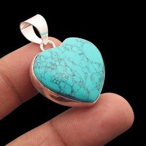 May include: A heart-shaped pendant with a turquoise stone and a silver-coloured bail. The turquoise stone has a marbled appearance with dark grey veins. The pendant is held by a finger against a black background.