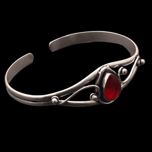 Puede incluir: Una pulsera de plata con una gema roja oscura en forma de lágrima. La pulsera presenta adornos en espiral y pequeños detalles redondos. El diseño abierto facilita su uso. La joya está sobre un fondo negro.