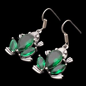 May include: Silver-coloured frog earrings with green gemstone detailing. Each earring has a frog design with a large, teardrop-shaped gem and smaller leaf-shaped gems. The background is black.