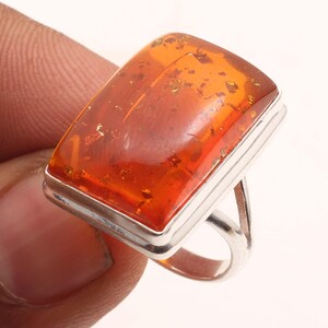 May include: A rectangular amber gemstone ring with a silver band. The amber stone has a warm orange hue with golden inclusions. The ring's band is a simple, polished silver design.