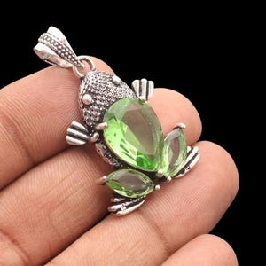 May include: A silver frog pendant with a large, teardrop-shaped green gemstone and smaller green gems. The frog's head is textured, and the pendant has a loop for a chain. The pendant is held in a hand.