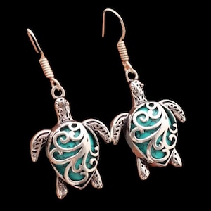 May include: A pair of silver-toned dangle earrings featuring a turtle-shaped charm. Each turtle has a decorative filigree metal overlay in a silver finish, set against a bright turquoise-colored inlay on the shell. The earrings hang from silver-toned hooks.