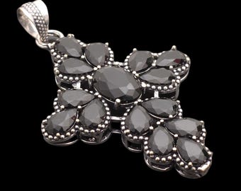 Puede incluir: Colgante de cruz plateada con un diseño detallado. La cruz está adornada con numerosas piedras preciosas negras de varias formas, incluyendo óvalos y lágrimas. El colgante tiene una argolla texturizada.