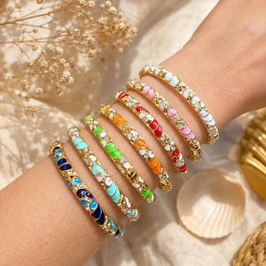 Op de afbeelding: Een set van zeven goudkleurige metalen armbanden, elk met een uniek emaille kleurpatroon in donkerblauw, lichtblauw, groen, oranje, rood, roze en wit, geaccentueerd met kleine heldere kristallen ingelegd langs de gehele buitenzijde.