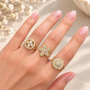 May include: Three gold-toned rings featuring clear, sparkling stones. One ring has a circular design with a four-leaf clover motif, the second is a five-pointed star shape, and the third is a sunflower-inspired design with layered petals.