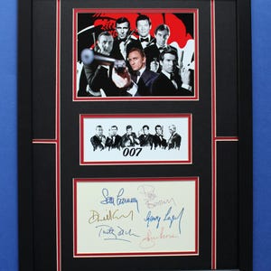 May include: A framed print featuring images of James Bond actors, including Daniel Craig, set against a red and black backdrop. The print includes a group photo and a section with signatures. The black frame has red borders.