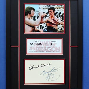 Peut inclure: Impression encadrée avec Chuck Norris et Bruce Lee dans une scène de "La Fureur du Dragon". L'impression comprend une photo des deux acteurs, une carte de titre et des signatures. Le cadre est noir avec un passe-partout rouge et blanc.