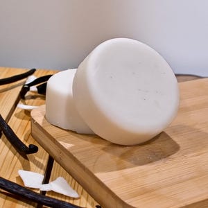 Puede incluir: Dos jabones redondos y blancos apilados sobre una pequeña tabla de cortar de madera. Vainas de vainilla y copos de coco esparcidos alrededor de los jabones. Los jabones tienen una superficie lisa y unas pocas motas oscuras.