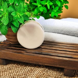 May include: A spa-themed still life featuring a round, off-white soap bar on a dark brown wooden soap dish. Fresh green mint plants in a small terracotta pot and folded white towels complete the scene. The setting is on a woven mat.