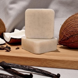 May include: Two square, light-colored soaps with dark specks are stacked on a wooden board. The soaps are surrounded by a coconut, coconut pieces, vanilla beans, and other ingredients, suggesting a natural skincare product.