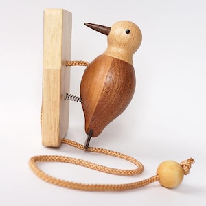 May include: A wooden woodpecker toy with a light brown head and a darker brown body. The bird is attached to a wooden block with a spring and a tan rope with a wooden ball at the end. The toy is designed to look like the bird is pecking.