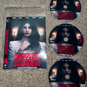 Puede incluir: Un set de DVD de la serie de Netflix "Something Very Bad Is Going to Happen: Complete Season One". La portada presenta a una mujer con cabello oscuro. También son visibles tres DVD, cada uno etiquetado como "Disco 1", "Disco 2" y "Disco 3".