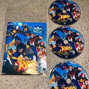 Puede incluir: Un set de DVD de la temporada 2 de X-Men '97, que incluye una funda rectangular y tres discos circulares. La ilustración muestra varios personajes con trajes azules y amarillos, con el logotipo de X-Men '97 en texto amarillo y rojo sobre fondo azul.