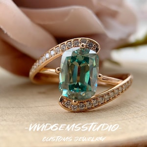 May include: A rose gold bypass ring featuring a central cushion-cut teal gemstone flanked by two curved bands encrusted with small white stones. The text VIVIDGEMSSTUDIO CUSTOMS JEWELRY is printed at the bottom of the image.