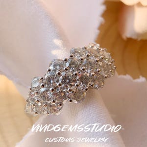 May include: A sparkling silver ring with numerous clear gemstones. The ring is set against a white fabric background, with the text "-VIVIDGEMSSTUDIO- CUSTOMS JEWELRY" at the bottom. The ring's design is clustered and eye-catching.